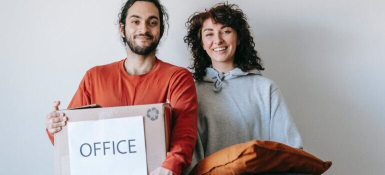 A man holding box labeled office and woman holding pillows 